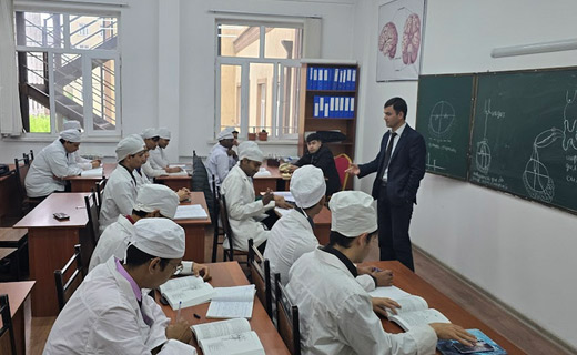 A classroom with a group of students in white lab coats taking notes while a teacher in a dark suit lectures and points to a chalkboard with anatomical drawings at Tajhind Edutech Pvt Ltd.
