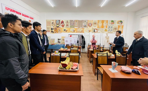 People standing in a classroom with various anatomical models and educational exhibits, including human organs and skeletal parts, while a person appears to be giving a presentation or lecture on MBBS in Tajikistan.