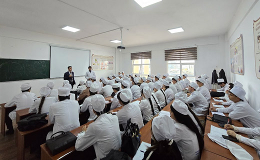 A classroom filled with students in white uniforms and head coverings, attentively listening to a teacher standing near a blackboard. There are educational posters on the walls promoting MBBS in Tajikistan.