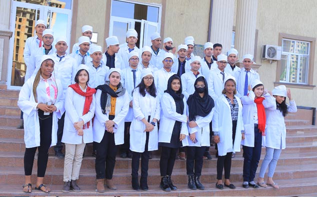 A group of medical students in white coats and caps posing on steps outside a building. some wear scarves and all appear focused and professional.