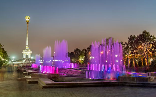 A nighttime view of a colorful illuminated fountain with purple and pink lights, in a park with a tall monument topped by a golden statue in the background.