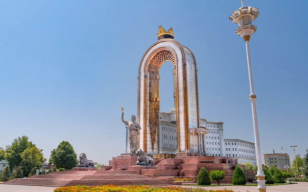 Image of the independence arch in tajikistan featuring a golden crown at the top, statues, and lions next to a large government building under a clear blue sky.