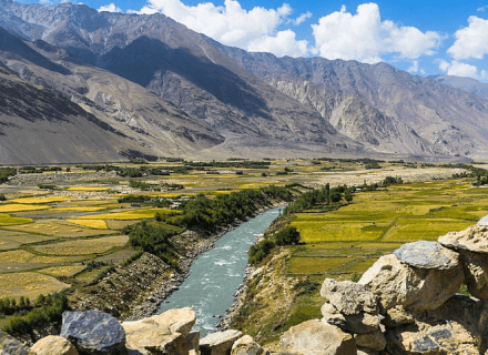 A scenic view of a vibrant blue river flowing through a lush valley with patchwork fields and surrounded by rugged mountains under a clear sky, captured during an educational trip organized by Tajhind Edutech Pvt