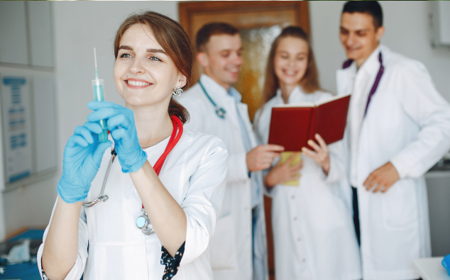 A female doctor in a lab coat and gloves holds a syringe, smiling confidently. In the background, three medical professionals discuss notes and smile, in a bright clinical setting while considering opportunities to
