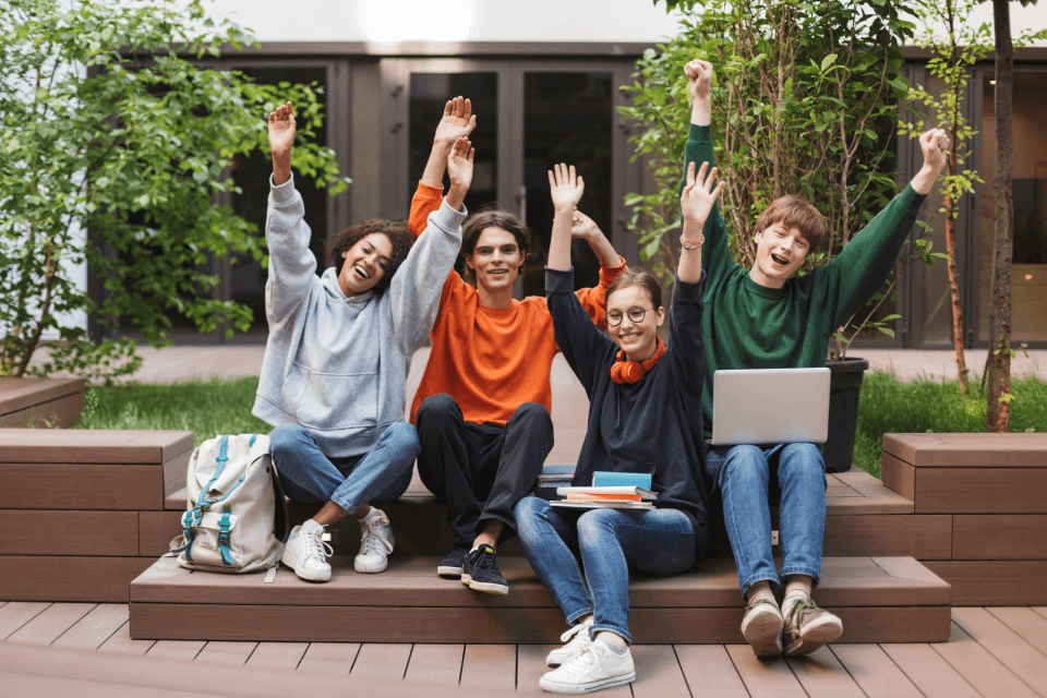 Four young students sitting on outdoor steps, joyfully raising their arms in celebration. They are diverse and casually dressed, with a laptop and books around them, discussing their MBBS studies in Tajikistan
