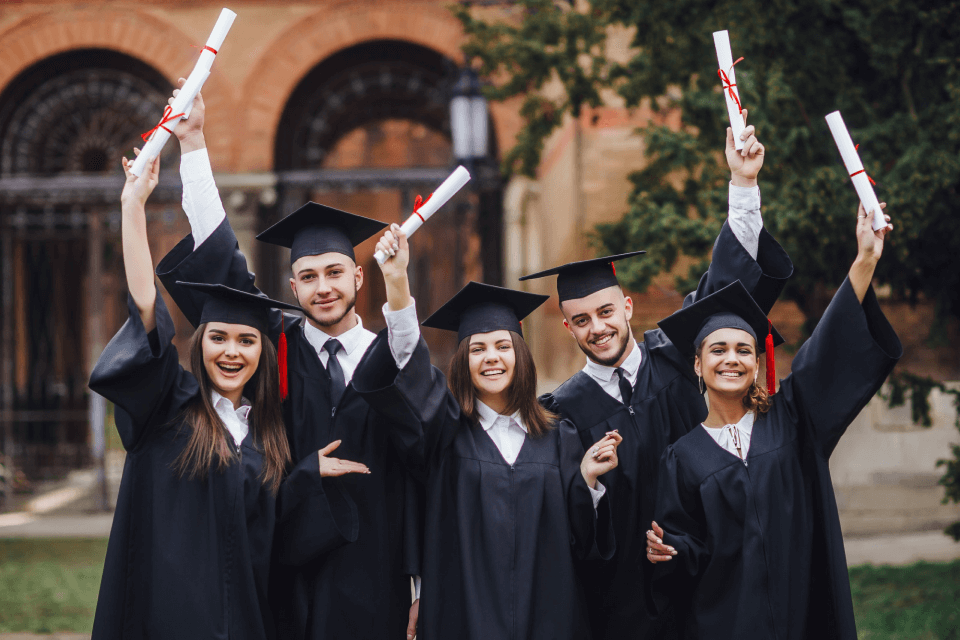 Five graduates in caps and gowns joyfully hold up their diplomas in front of a brick building, smiling and celebrating their achievement.