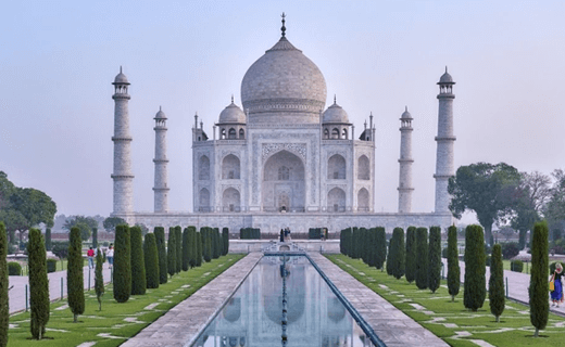 View of the taj mahal at dawn, showing its iconic white marble dome and minarets, reflected in the long, narrow pool surrounded by manicured gardens.