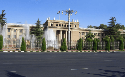 An elegant historic building surrounded by manicured gardens, fountains, and ornate street lamps, viewed from across a wide, empty street.