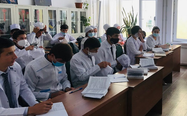 Medical students in white coats and face masks sit at desks in a classroom, taking notes from textbooks during a lecture on Study MBBS Abroad.