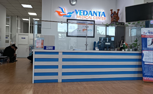 Interior of Vedanta Medical Center with a reception desk showcasing blue and white colors, various informational posters about MBBS in Tajikistan, and a person cleaning the floor on the left.