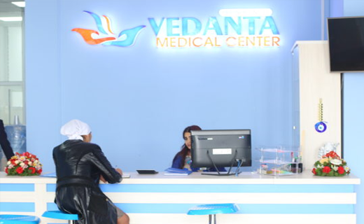 A patient speaks with a receptionist at Vedanta Medical Center's front desk, which features a blue and orange logo. The interior is modern with floral decorations and promotional material for Tajhind Edutech